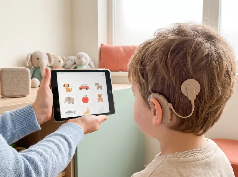 In einem hellen Therapieraum hält eine Fachkraft ein Tablet mit Bildkarten, ein Kind greift mit der Hand danach