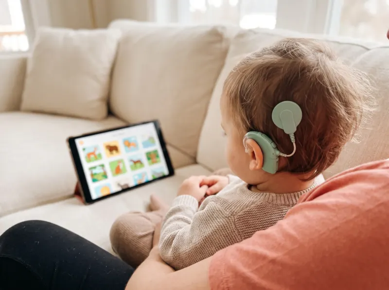 Eltern und Kind spielen gemeinsam auf dem Sofa ein Hörspiel am Tablet, das Kind trägt einen Cochlea-Implantat-Prozessor und tippt auf eine Bildkarte